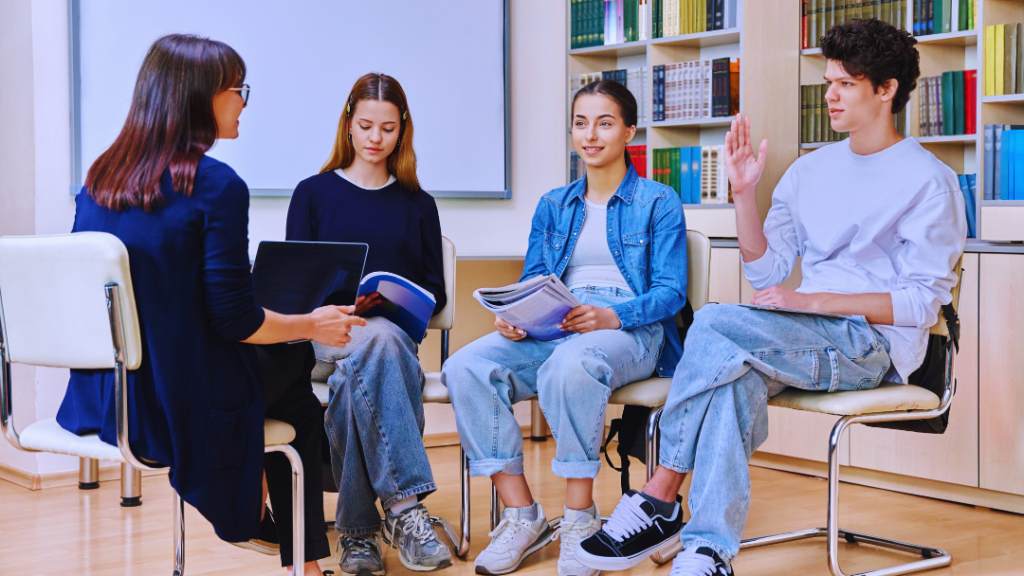 Youth worker in blue cardigan facilitates discussion with 3 diverse teens holding books & laptop in library circle. One boy raises hand amid bookshelves & whiteboard. Promotes engagement & learning.