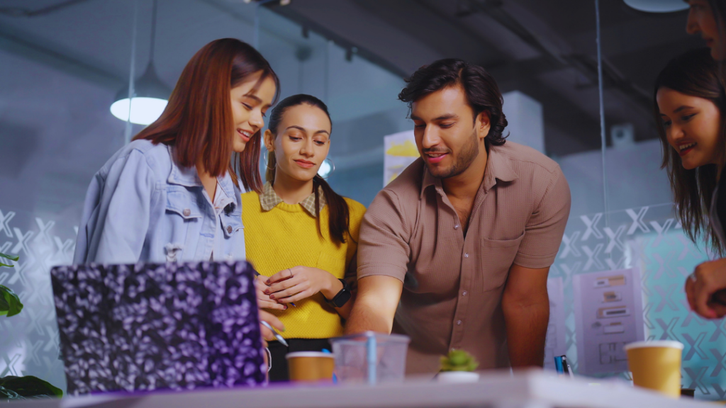 A group of four young professionals, three women and one man, gather around a laptop and desk, smiling and collaborating. The Youth Workers are reviewing a project, suggesting teamwork and mentorship.